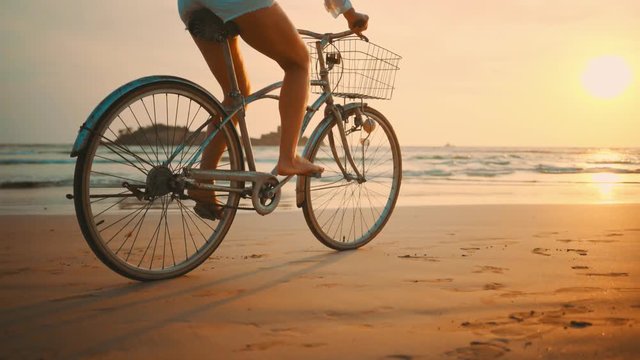 Woman in white and hat mooving on her bicycle along beach sand at summer time under burning sunset sky, lifestyle carefree.