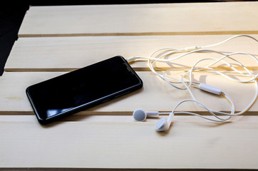 Black smartphone with white headphone is lying on wooden background. Modern technology connection. Mobile phone and earphone digital cell.
