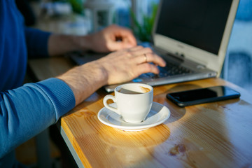 Close-up of men's hands, typing on a laptop, drinking coffee, next to the phone. A cup of coffee is empty. Close-up. Sunlight