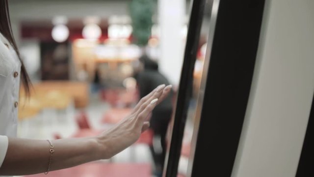 Caucasian female using automated teller machine with big digital screen while standing in shopping mall, woman verifies account balance on banking application via modern device icon