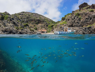 Obraz premium Boat moored near a cove with a fishermen hut and a shoal of fish underwater, split view above and below water surface, Cap de Creus, Mediterranean sea, Spain, Costa Brava, Catalonia, Cala Gentil