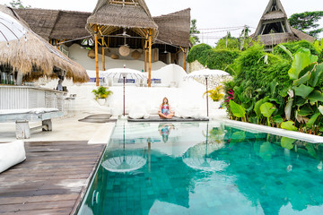 A young girl in a swimsuit is sitting in a yoga pose near the pool, in the background there are greens and palms. Luxury hotel with bungalow. Everything is white and bright. View with copy space