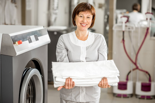 Portrait of a senior washwoman in uniform standing with bedclothes in the hotel laundry