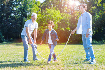 Positive vibes. Joyful familys men generation grinning broadly into the camera while spending their free time outdoors and playing with a jump rope in the park.