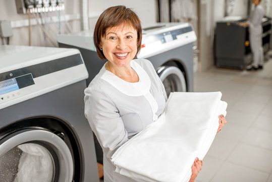 Portrait of a senior washwoman in uniform standing with bedclothes in the hotel laundry