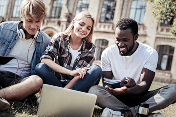 International groupmates. Delighted happy positive student sitting together on the grass and looking at the laptop screen while preparing home task together