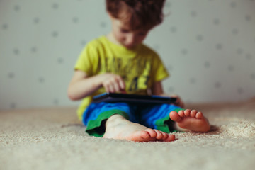 Kid with tablet sitting on bed and watching cartoons. Selective Focus on the feet. Cute boy in green t-shirt and blue pants