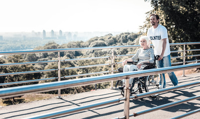 People with disability. Cheerful positive handsome man standing behind the wheelchair and pulling it while helping elderly people