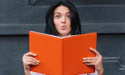 Beautiful and funny female student holds book, has shocked expression as tomorrow is exam, on black background. Studying, education and business concept
