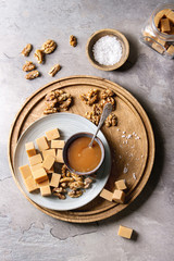 Salted caramel fudge candy served on wooden board with fleur de sel, caramel sauce and caramelized walnuts in bowls over grey table. Top view, space. Dessert set