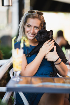 Beautiful young blond woman with sunglasses enjoying with her french bulldog puppy in cafe bar.