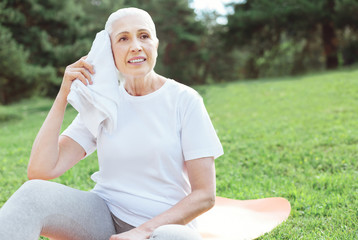 Fototapeta premium Minute of rest. Positive nice elderly woman sitting on the yoga mat and holing a towel while having a rest during a workout
