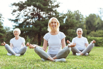 Yoga class. Delighted positive aged woman sitting on the grass and smiling while practicing yoga with her friends