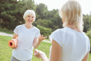 Pleasant communication. Happy delighted senior woman smiling and talking to her friend while holding a yoga mat