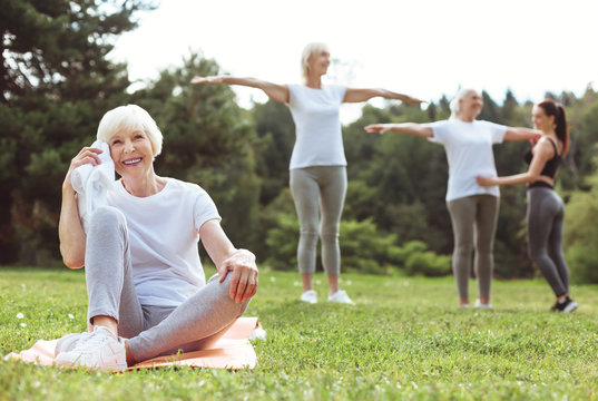 Positive mood. Cheerful joyful elderly woman sitting on the yoga mat and holding the towel while resting after a workout