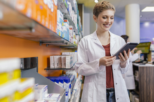 Smiling pharmacist at shelf with medicine in pharmacy holding tablet