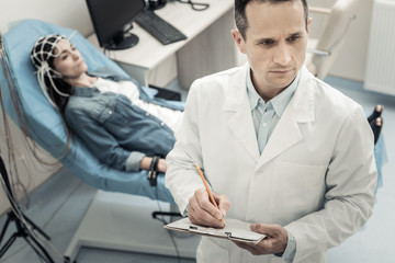 Doctors notes. Serious nice skillful doctor holding a pencil an taking notes while standing in his office