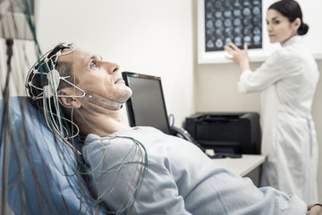 Electro scanning. Nice pleasant handsome man lying on the medical bed and wearing electronic wires while having his brain scanned - obrazy, fototapety, plakaty