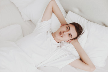 Obraz na płótnie Canvas A handsome young man lies on a bed and smiles thoughtfully, putting his hands behind his head. Dream about the future. White bed.