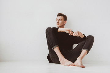 A young man, a student sits on the floor, against a white wall background, a pensive look. The guy...