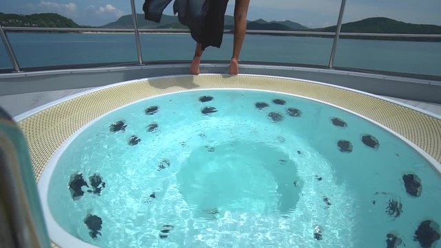 Asian girl in a pink swimsuit and black cloak posing near jacuzzi on a luxury yacht. Hot tub jacuzzi with turquoise water on the boat.