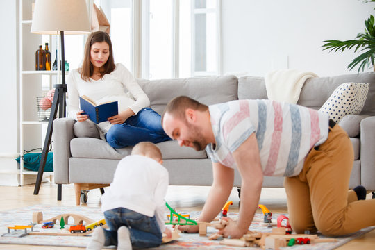 Family photo of father and son playing on floor in toy road on background of pregnant woman reading book lying on couch
