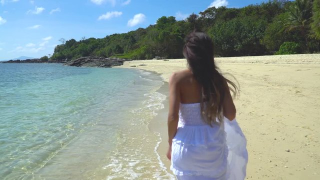 A girl in a white dress is runing along the shore on the white sand beach near the turquoise sea water. View from the back. Thailand.
