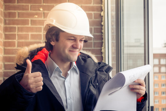 Handsome architect is standing near building outdoors. He is giving thumb up and smiling. The man in helmet is looking at camera happily. Soft focus, toned.