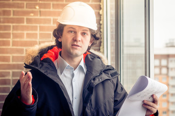 An angry disgruntled builder worker in a helmet with project drawings plans in his one hand and mobile phone in another hand screams on someone. Bad executed work. Aggressive man. Soft focus, toned.