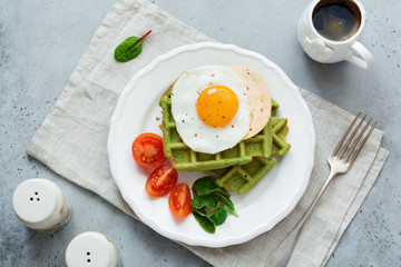 Fried egg with cheese, tomatoes, chard, and waffle with spinach on white ceramic plate on light gray concrete background. Sandwich for breakfast. Selective focus. Top view. Copy space.