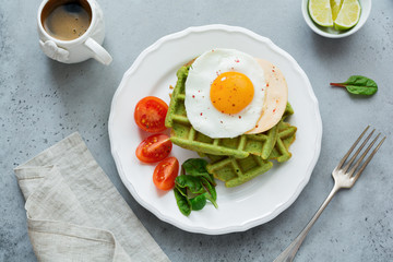 Fried egg with cheese, tomatoes, chard, and waffle with spinach on white ceramic plate on light gray concrete background. Sandwich for breakfast. Selective focus. Top view. Copy space.