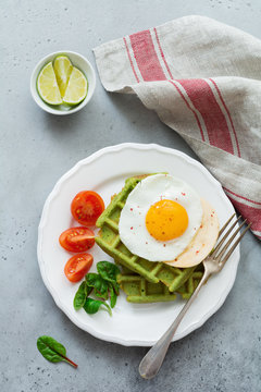 Fried egg with cheese, tomatoes, chard, and waffle with spinach on white ceramic plate on light gray concrete background. Sandwich for breakfast. Selective focus. Top view. Copy space.