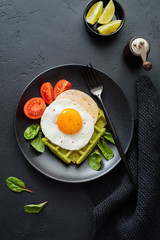 Fried egg with cheese, tomatoes, chard, and waffle with spinach on black ceramic plate on dark concrete background. Sandwich for breakfast. Selective focus. Top view. Copy space.