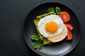 Fried egg with cheese, tomatoes, chard, and waffle with spinach on black ceramic plate on dark concrete background. Sandwich for breakfast. Selective focus. Top view. Copy space.
