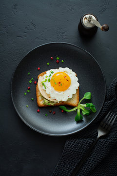 Toast with guacamole sauce from avocado, cheese, fried egg and fresh on black ceramic plate on dark concrete background. Selective focus. Top view.