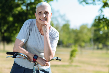 Positive mood. Delighted cheerful elderly woman holding her chin and looking at you while standing with a bicycle
