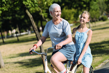 Obraz premium Such a pleasure. Cheerful positive elderly woman sitting on the bike and smiling while enjoying her ride together with the granddaughter