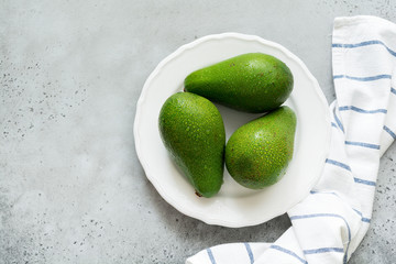 Three raw fresh avocados in white old ceramic plate on gray concrete background. Selective focus. Top view.
