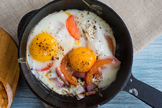 Fried eggs with tomatoes and onions in a cast iron pan on a rustic wooden background