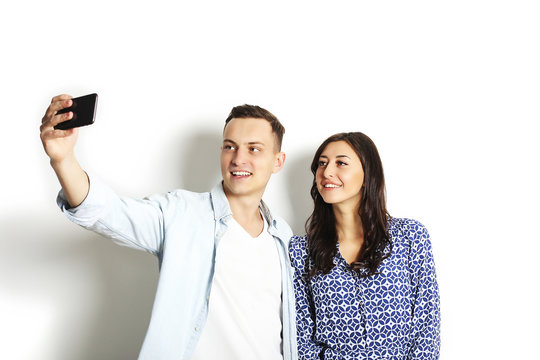 Beautiful excited young straight couple making funny faces, laughing, smiling & taking selfie, having fun w/ mobile cell phone camera gadget. Portrait, copy space, isolated white wall background.