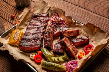 Grilled pork ribs served with grilled corn, salat, bbq sauce, salt pepper and cucumber on parchment paper on a wooden table. Top View
