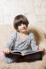 A little smiling girl with blue eyes is reading a book.