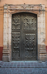 An old elaborate door, with beautifully crafted details and a massive stone frame, with a brick walkway, in Guanajuato, Mexico - 191377182
