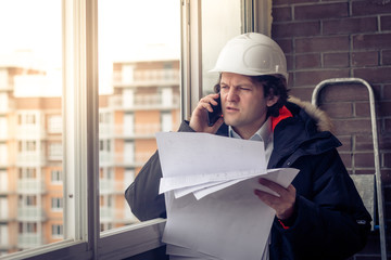 Portrait of concerned unshaven builder telling by mobile while keeping different projects in hand....
