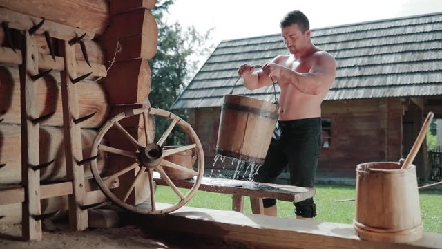 Attractive muscular shirtless young man carries the wooden bucket with water, puts it on the bench and freshens up in front of the wooden building. Crafts, traditions. Summertime, healthy lifestyle