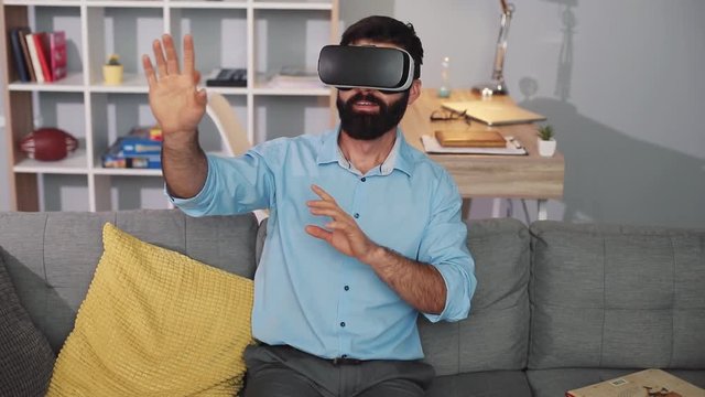 Close up handsome man wearing virtual reality glasses waving his hands sit on the sofa at home feel shock innovation looking modern entertainment gadget new tech visual beard game hipster slow motion