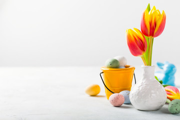 Easter composition with spring tulips flowers in white vase and colored quail eggs candies in bucket. Holidays concept with copy space.