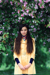 Spring portrait of a pretty girl with long dark curly hair under blooming apple tree with pink flowers on branches, Woman in yellow coat and bright scarf.