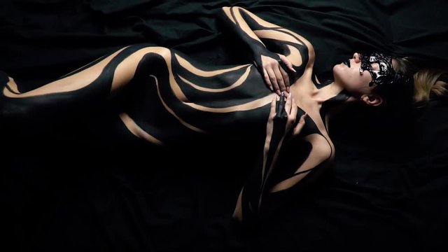 A girl in a mask and black body painting lay on a black silk sheet and covers her chest with her hands