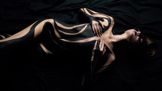 A girl in a lace mask and black body art lies on a black sheet
